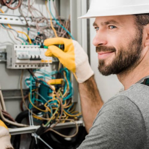 handsome cheerful electrician repairing electrical box and using screwdriver in corridor